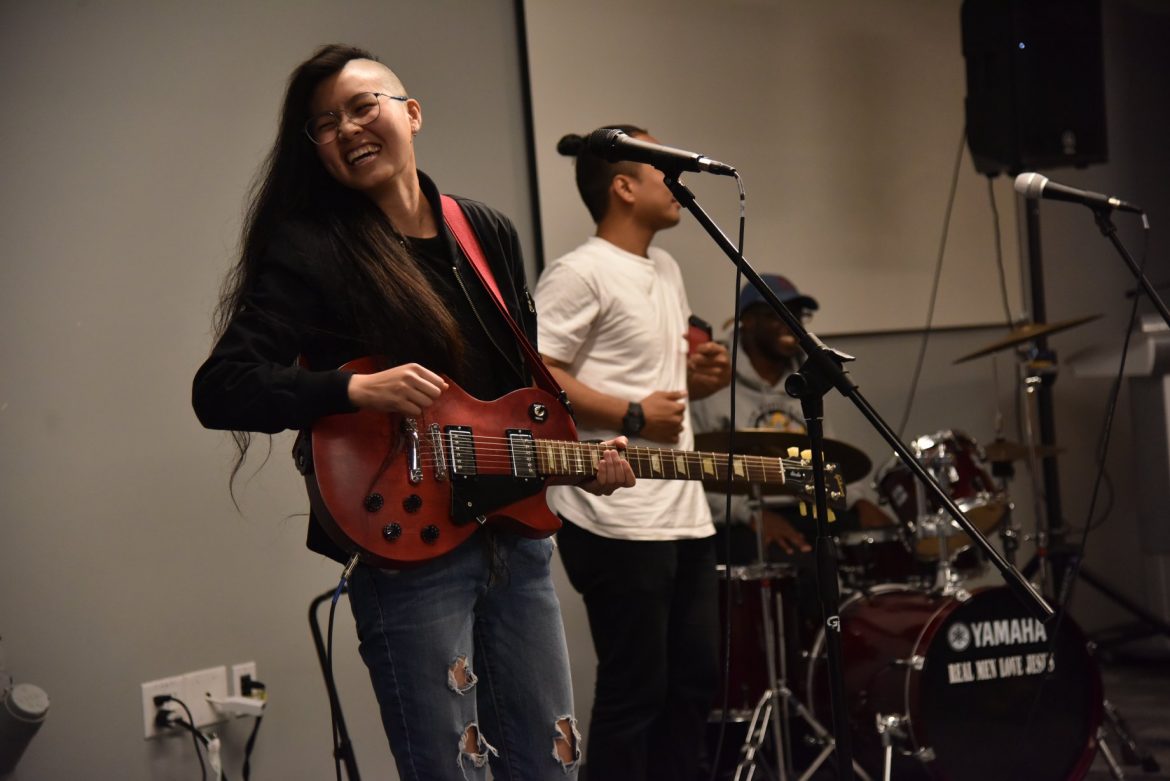 Image description: Yiann is standing while holding an electric guitar, smiling toward something to the right. (Photo credit: Corky Lee)