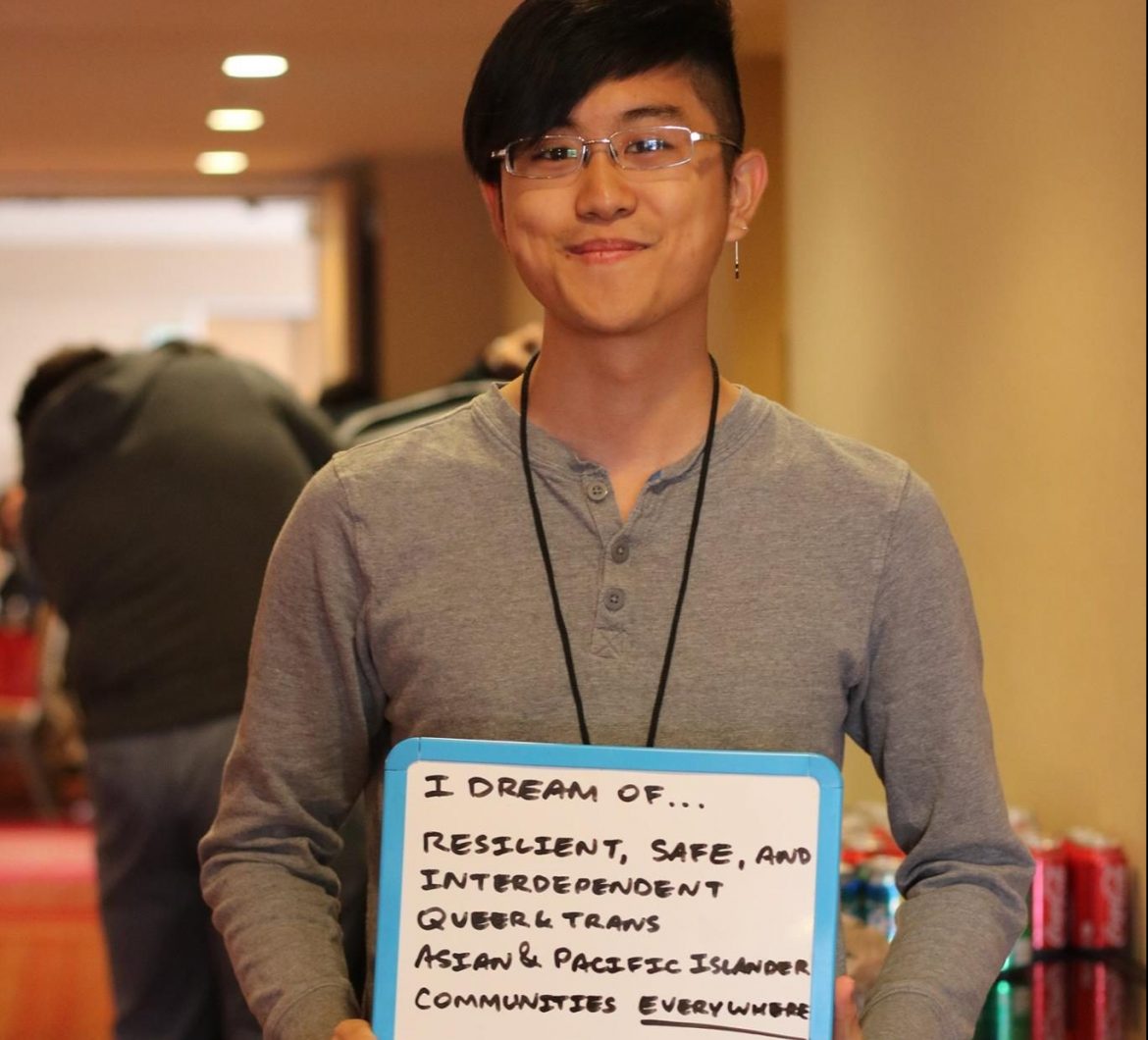 Image description: Ethan stands indoors, smiling at the camera and holding a small whiteboard that reads: "I dream of resilient, safe, and interdependent queer & trans Asian & Pacific Islander communities everywhere."