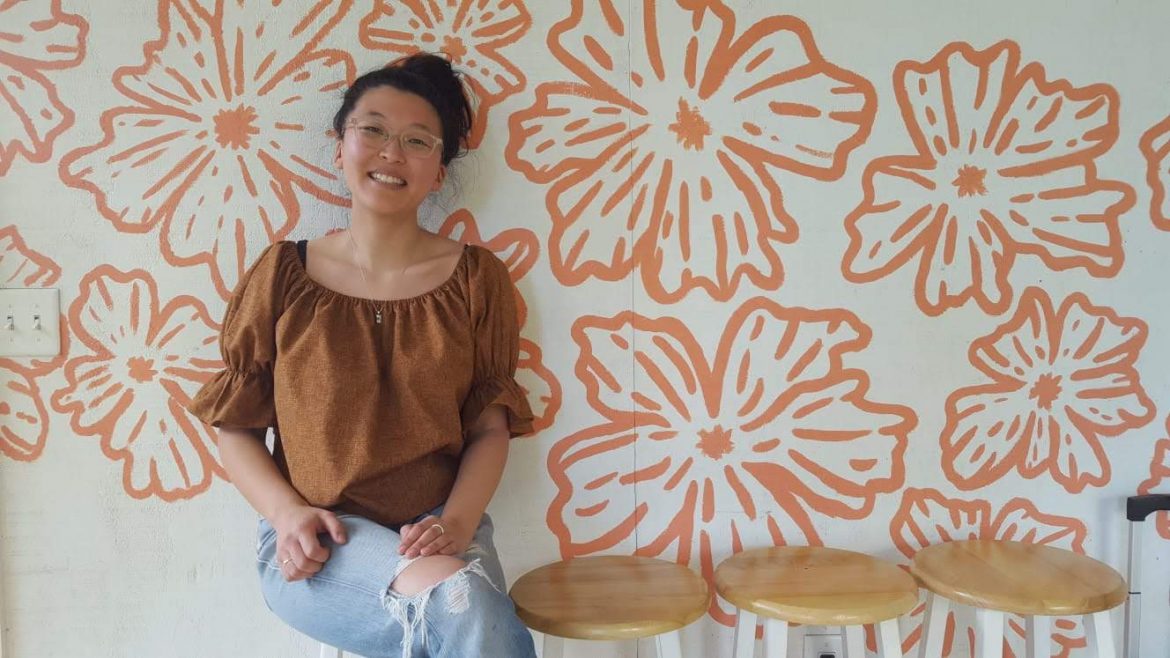 Mika sits indoors, smiling toward the camera. Behind her is a wall with big flowers prints.