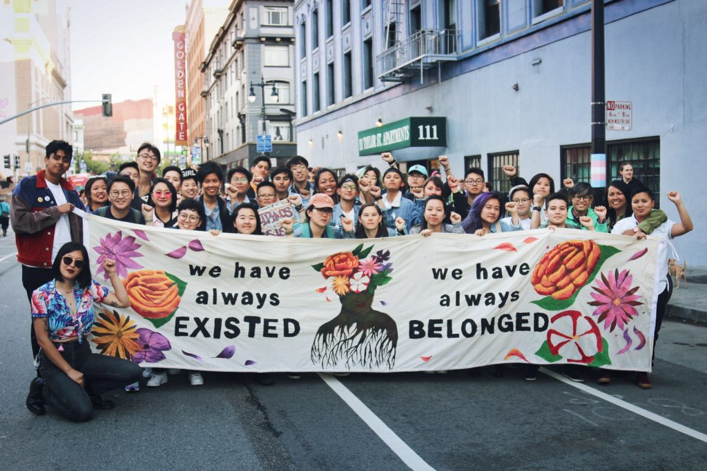 Image description: a large group of TGNC API people stand in the middle of the street with fists raised, holding banner that says "We have always existed; we have always belonged".