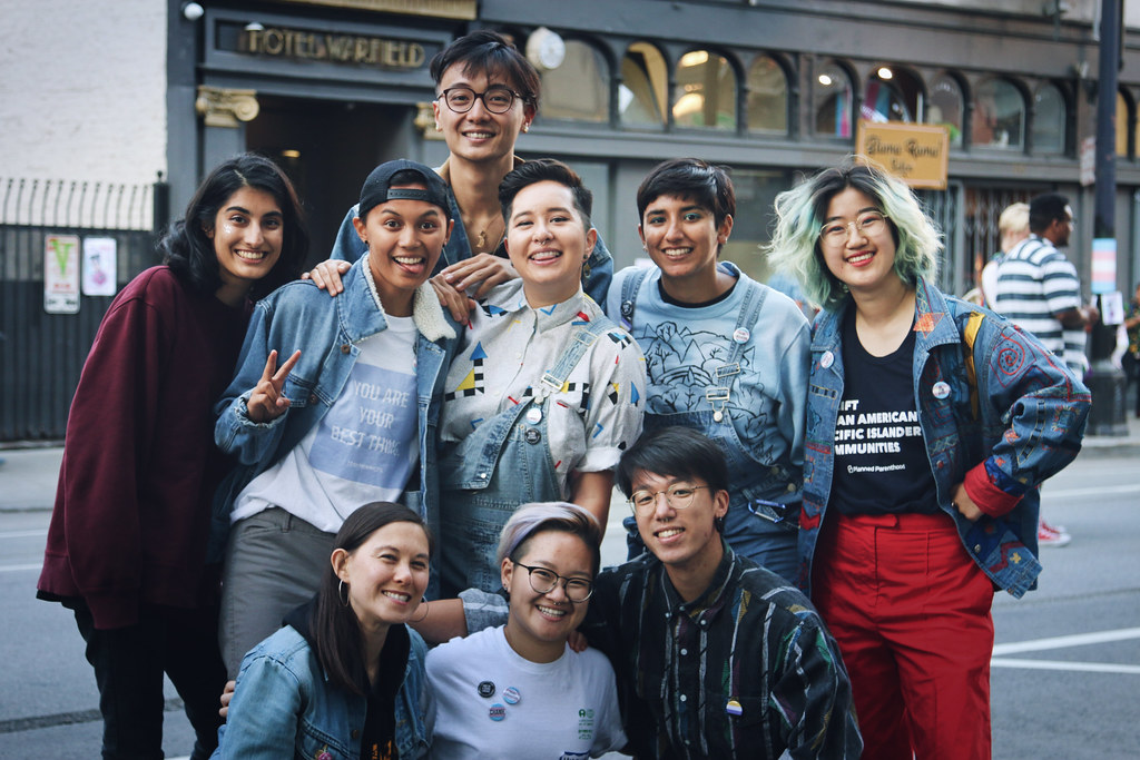 Image description: group photo of nine queer and tgnc API people kneeling and standing in the middle of the street, smiling toward the camera.