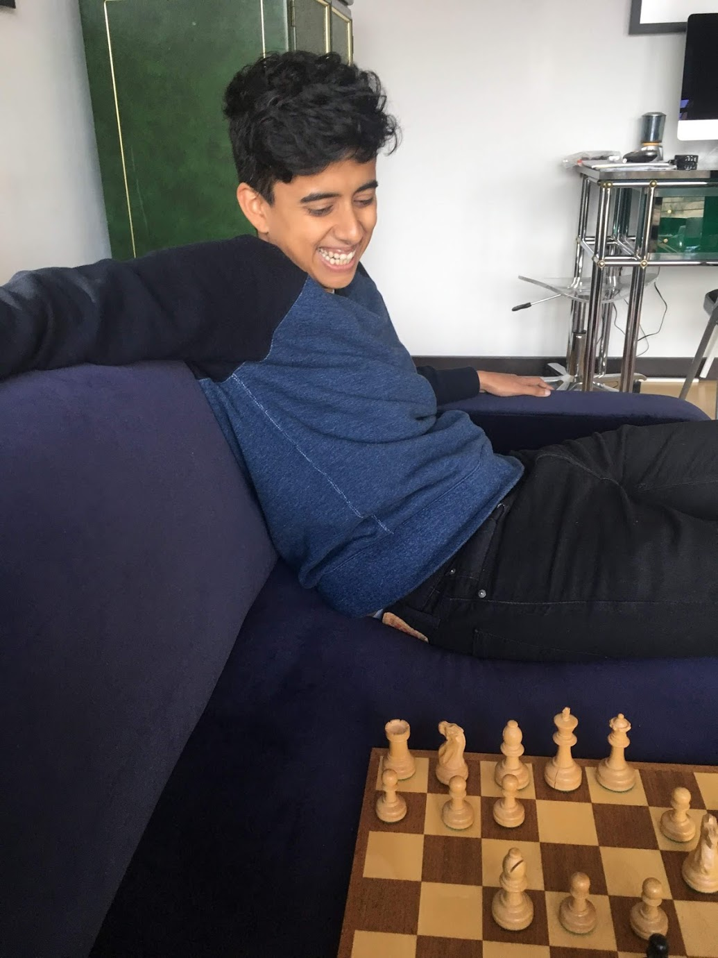 Image description: T is indoors, sitting on a purple sofa, right arm resting on the back of the sofa, looking down and smiling toward a chessboard.