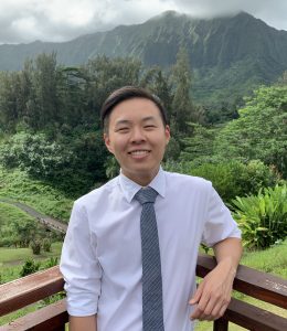 Image description: Kyle is outdoors and smiling on a balcony overlooking a lush forest and a mountain in the background.
