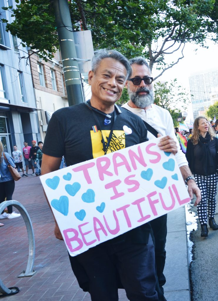 Image description: Vince stands on the sidewalk, smiling and holding a sign that says "Trans is Beautiful" in pink lettering and surrounded blue hearts.