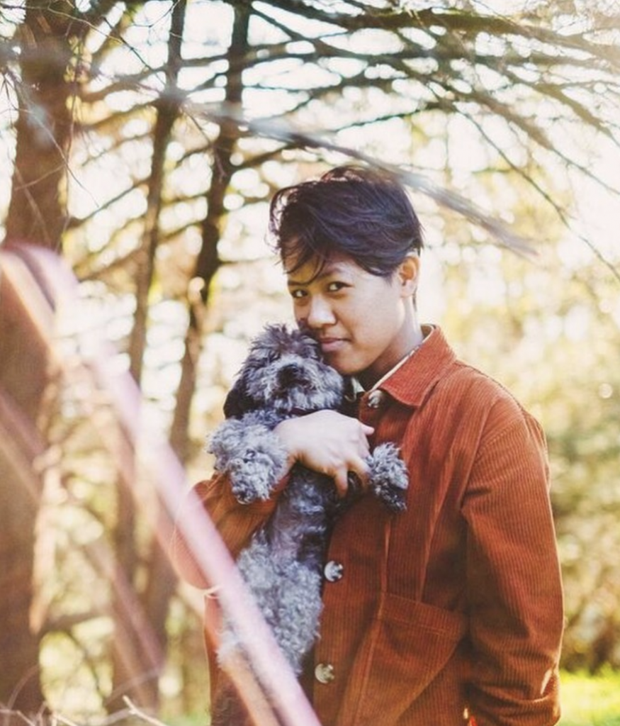 Image description: Gem stands outdoors amongst trees holding a fluffy gray dog in their arm while slyly smiling at the camera.