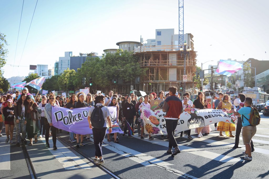 Image description: A large group of TGNC API people march up Market St. while holding banners, signs, and trans flags at 2019 SF Trans March. T is in the center, walking backwards.