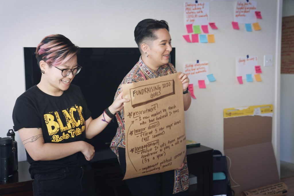 Image description: Huanvy and Sammie are smiling and holding up a flipchart indoors. There are papers and Post-It notes on the wall behind them.