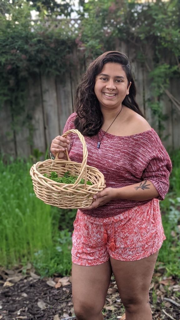Image description: Sangeeta is outdoors smiling while holding up a basket of plants. There is a fence and vegetation behind them.