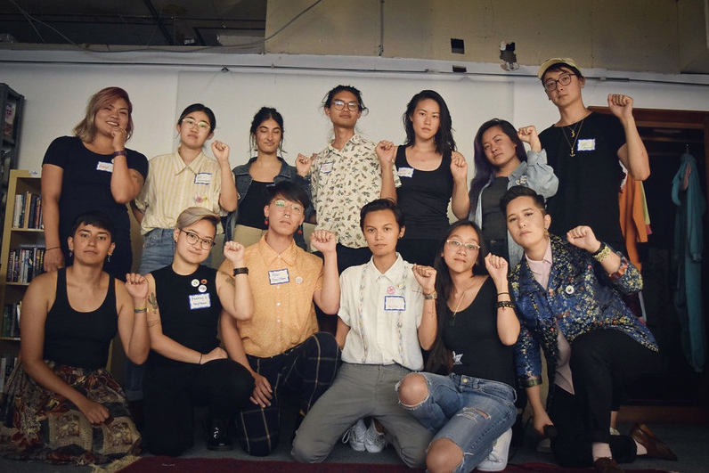Image Description: a group of 13 trans and queer API people, most in dark clothing, face the camera with serious faces and left fists raised in the air. Yuan is on the far right in the back row.