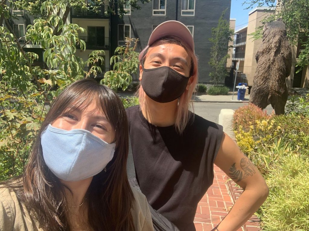 Image Description: Lily and Yuan face the camera smiling under face-masks. Surrounded by greenery.