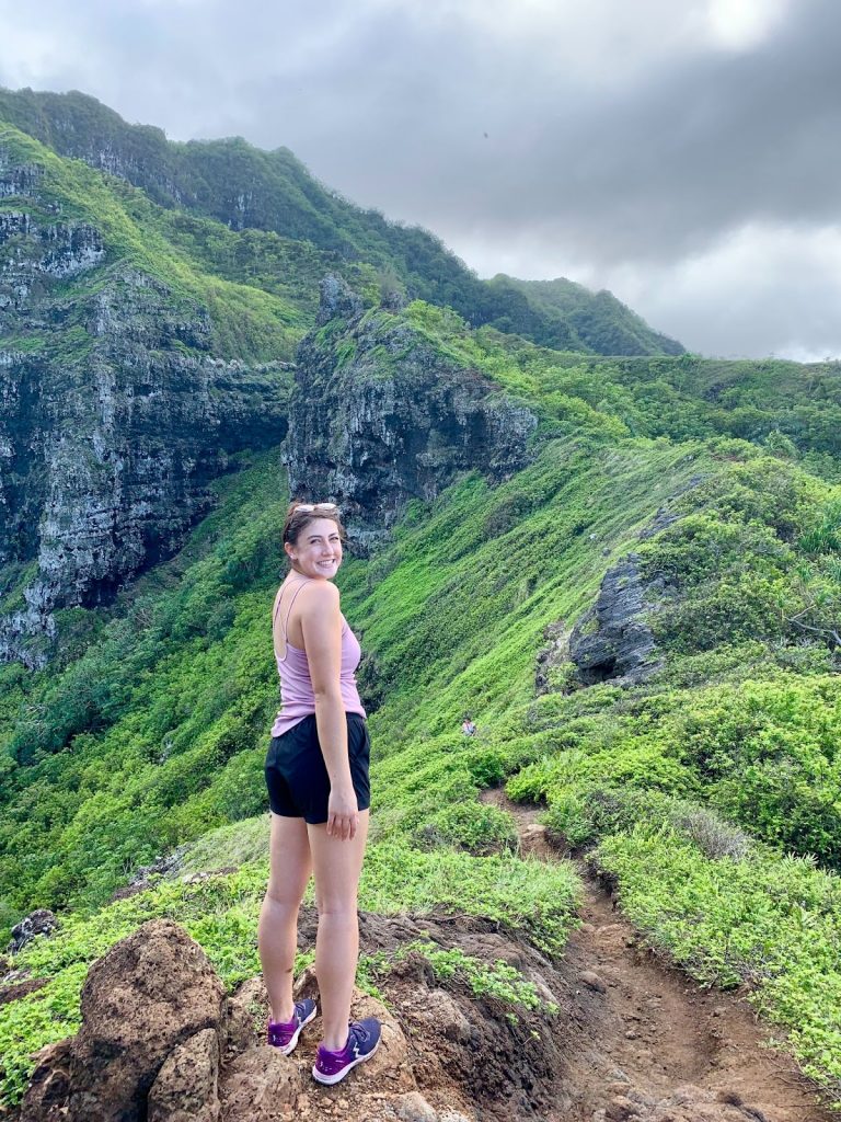 Image Description: Lily stands in a pink top and black shorts, turning back to the camera. Around her is a lush green hillside.