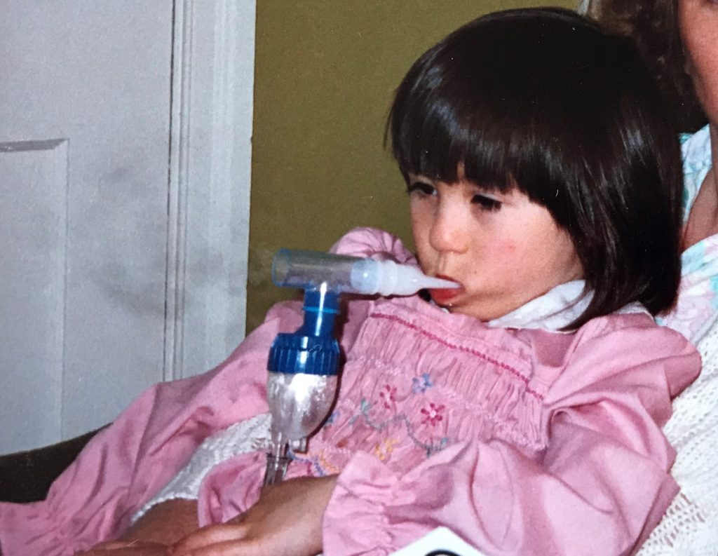 Image description: Photograph of Jasmin age 6 years old in a pink dress, taking asthma medication from a ventilator, looking sullen.