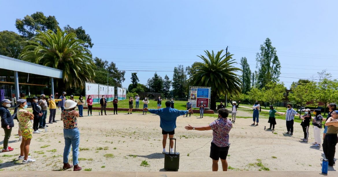 Image description: About 30 Lavender Phoenix members and staff standing in a half circle, with arms outstretched in a field at Peralta Park in Oakland. Shivani (they/them) is standing in the middle of the circle.