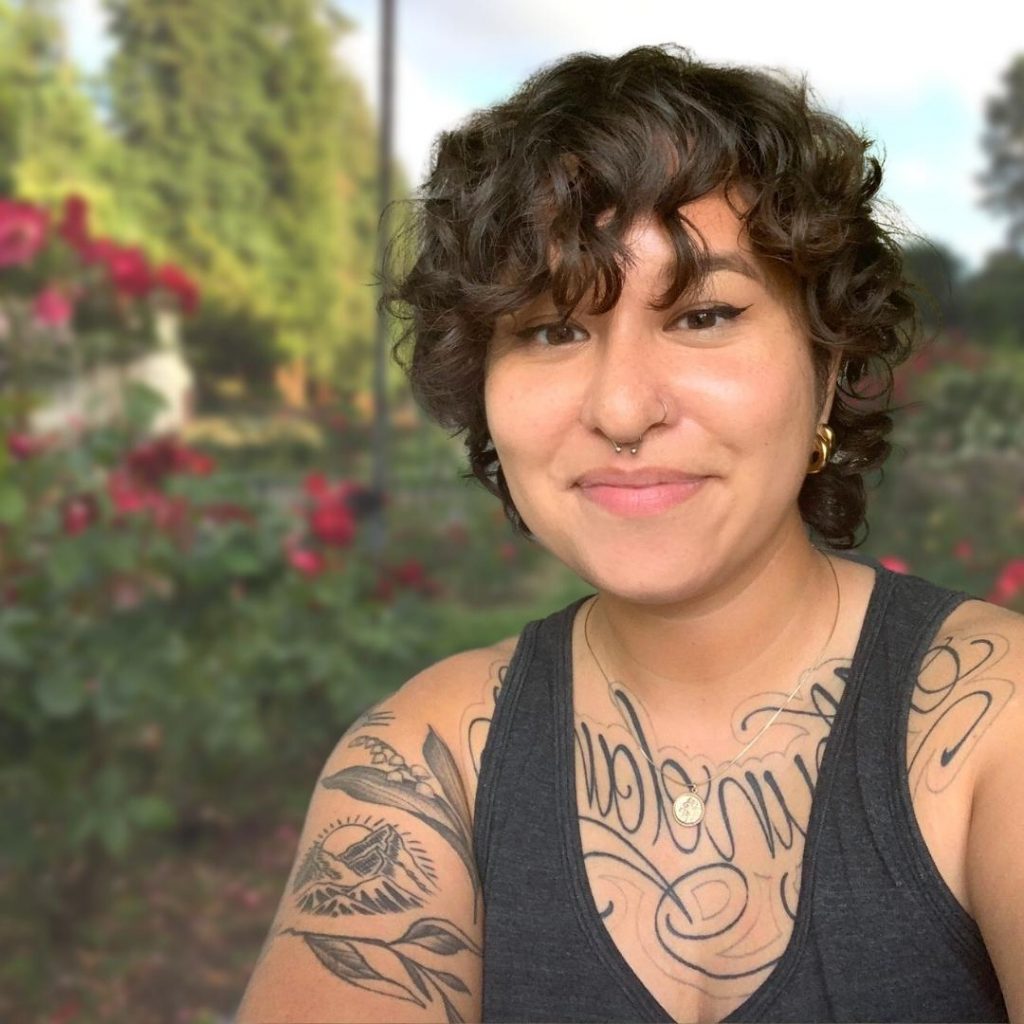 Image description: Sammie is in an outdoor garden setting smiling at the camera. They have curly hair and are wearing a tank top.