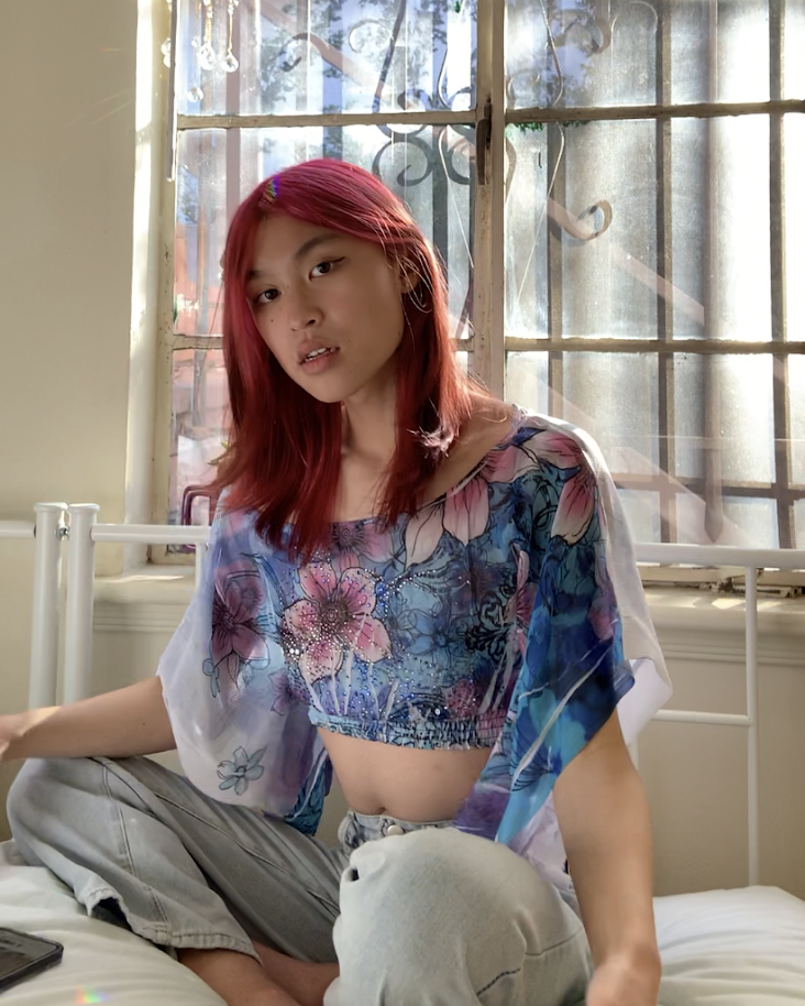image description: Iris (she/they), sitting on a white bed, with pink hair, softly looking at the camera. She is weathering a blue floral top and beige paints, sitting criss cross.
