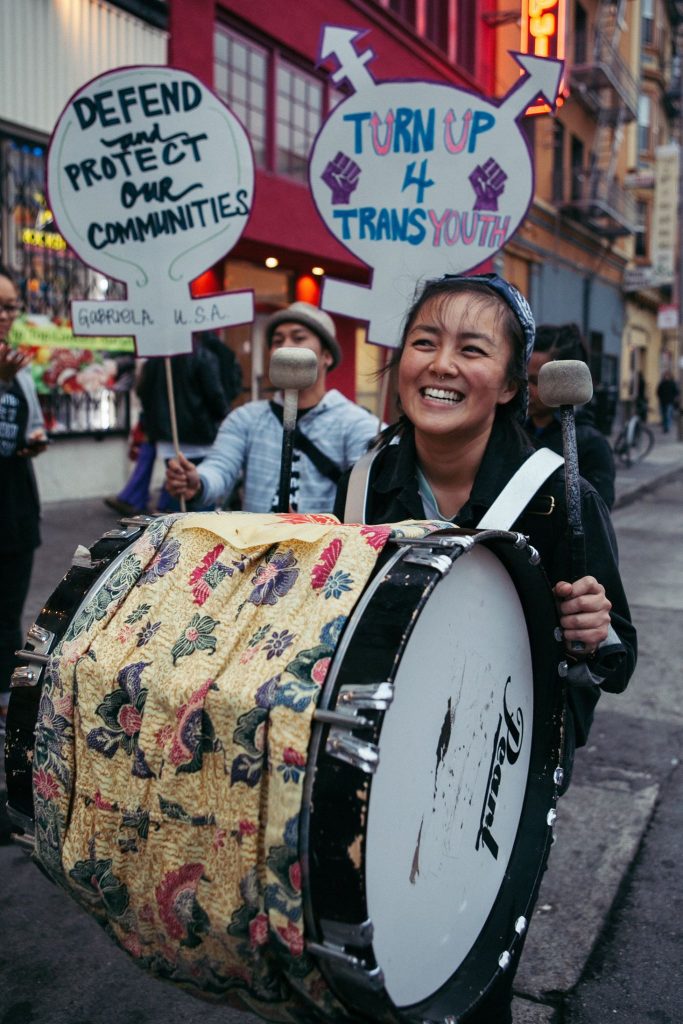 Image description: Cynthia smiles while playing a large drum strapped to their shoulders and marching.