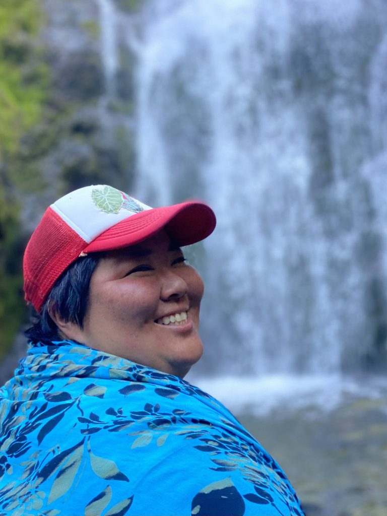 Image description: Eri in a blue outfit and a red cap smiles at the camera. There is a waterfall in the background.