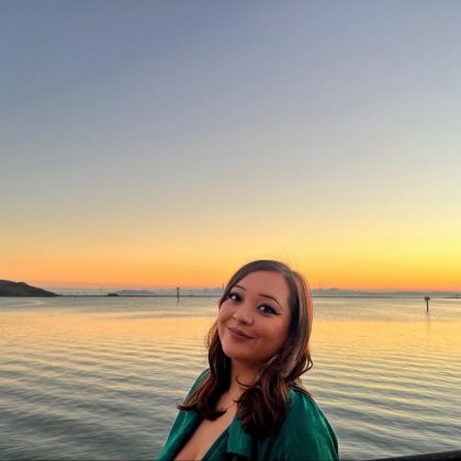 Image: Julz in a green top smiles at the camera. The sun sets over the water behind them.