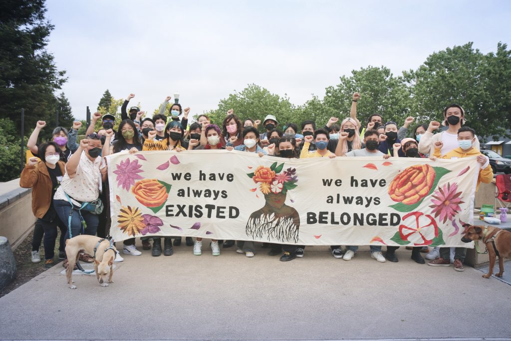 A crowd of trans/queer APIs raising fists behind a banner that reads “We have always existed, we have always belonged.” On the banner are a variety of hand-painted flowers.