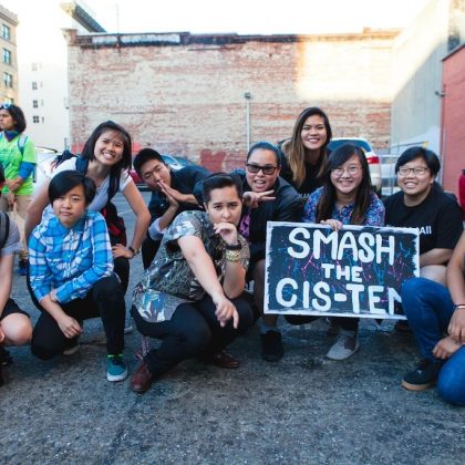 Image Description: 11 trans and queer APIs pose together at Trans March; one is holding a sign that reads: Smash the Cis-tem!