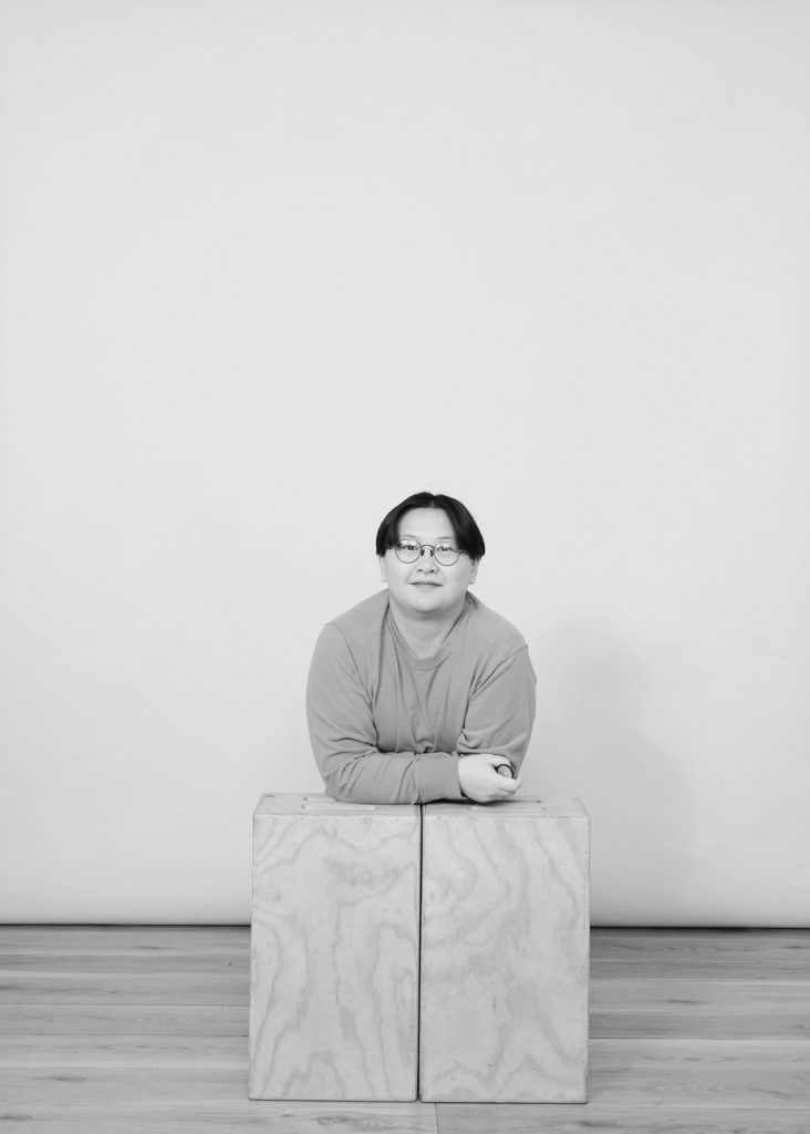 Image Description: a black and white image of Connie, who is posing leaning over two wooden blocks. Connie is smiling, wearing glasses and a long sleeve shirt.