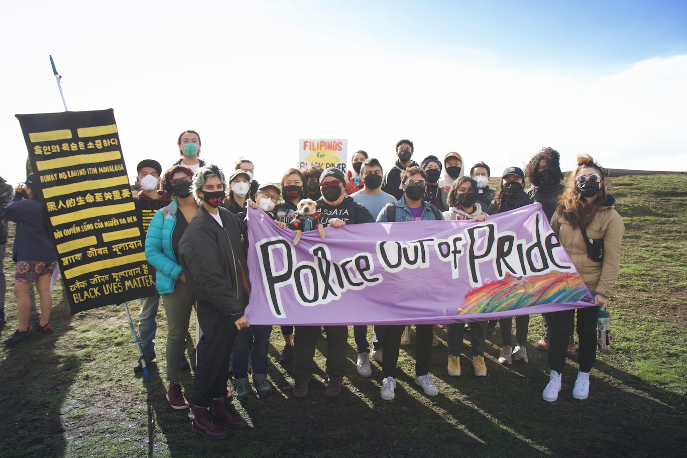 Image Description: A group of masked trans and queer Asian and Pacific Islander people hold a large purple banner that reads: Police Out of Pride. There is a black banner to the left that reads BLACK LIVES MATTER in multiple different languages.