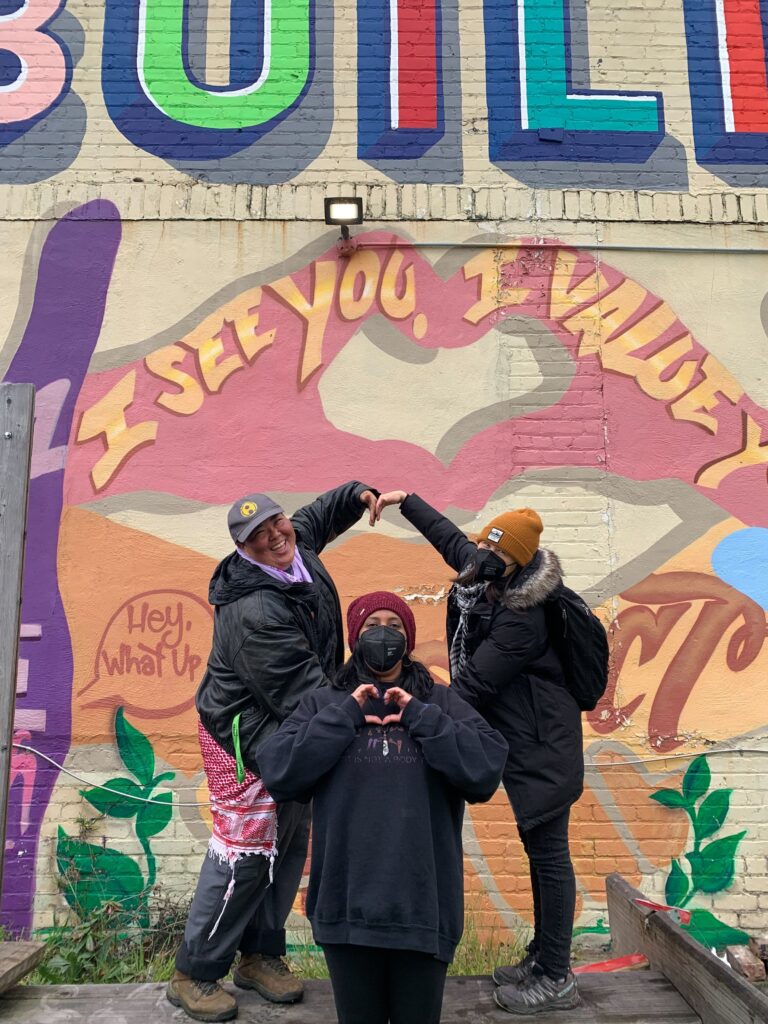 Image description: Three trans and queer API people in front of a brightly colored mural, collectively creating heart shapes with their arms and hands.