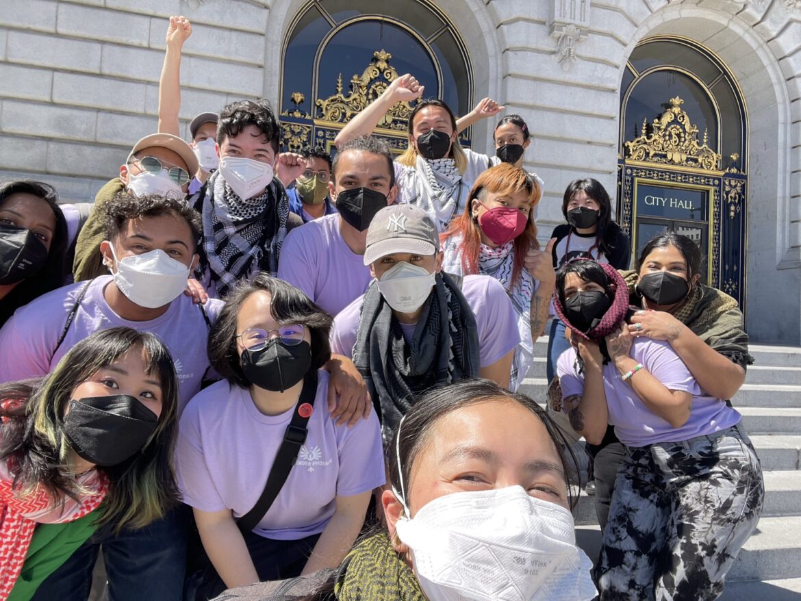 Image description: A group of trans and queer API people, many wearing lavender shirts and keffiyehs, gathered on the steps of San Francisco City Hall.