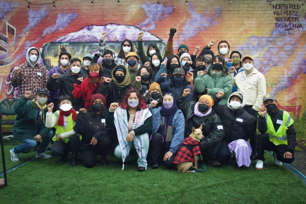 Image Description: A group of trans & queer API people standing and crouching in front of a mural. They are all masked and look fiercely towards the camera, some raising their fists.