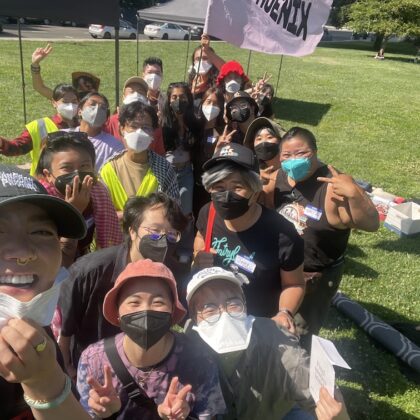 Image description: a group of ~20 masked Trans & Queer API people pose together for a selfie