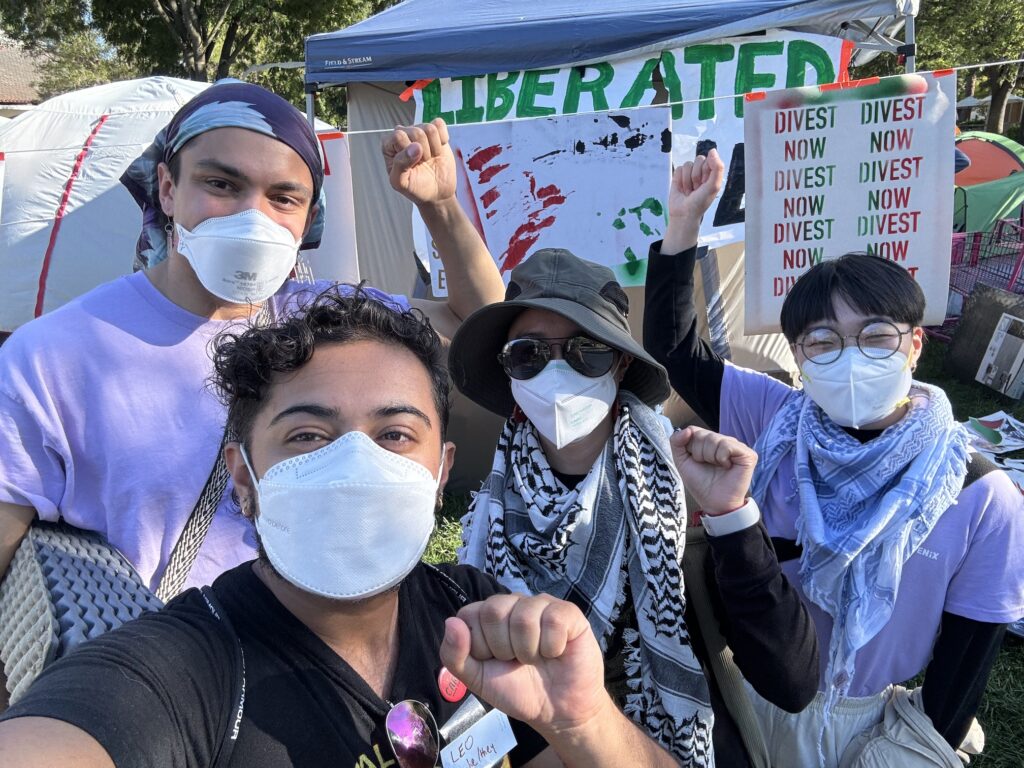 Image description: a group of four masked QTAPI people with fists raised in front of signs and tents at a student encampment for Palestine