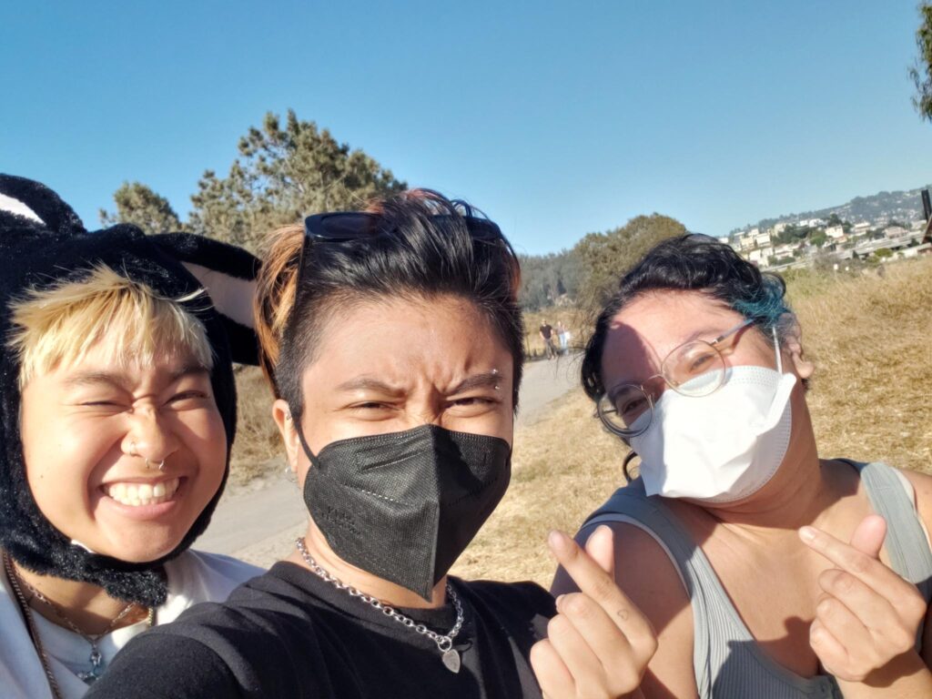 Image Description: 3 trans and queer Filipinos pose for the camera. 2 of them hold up finger hearts.