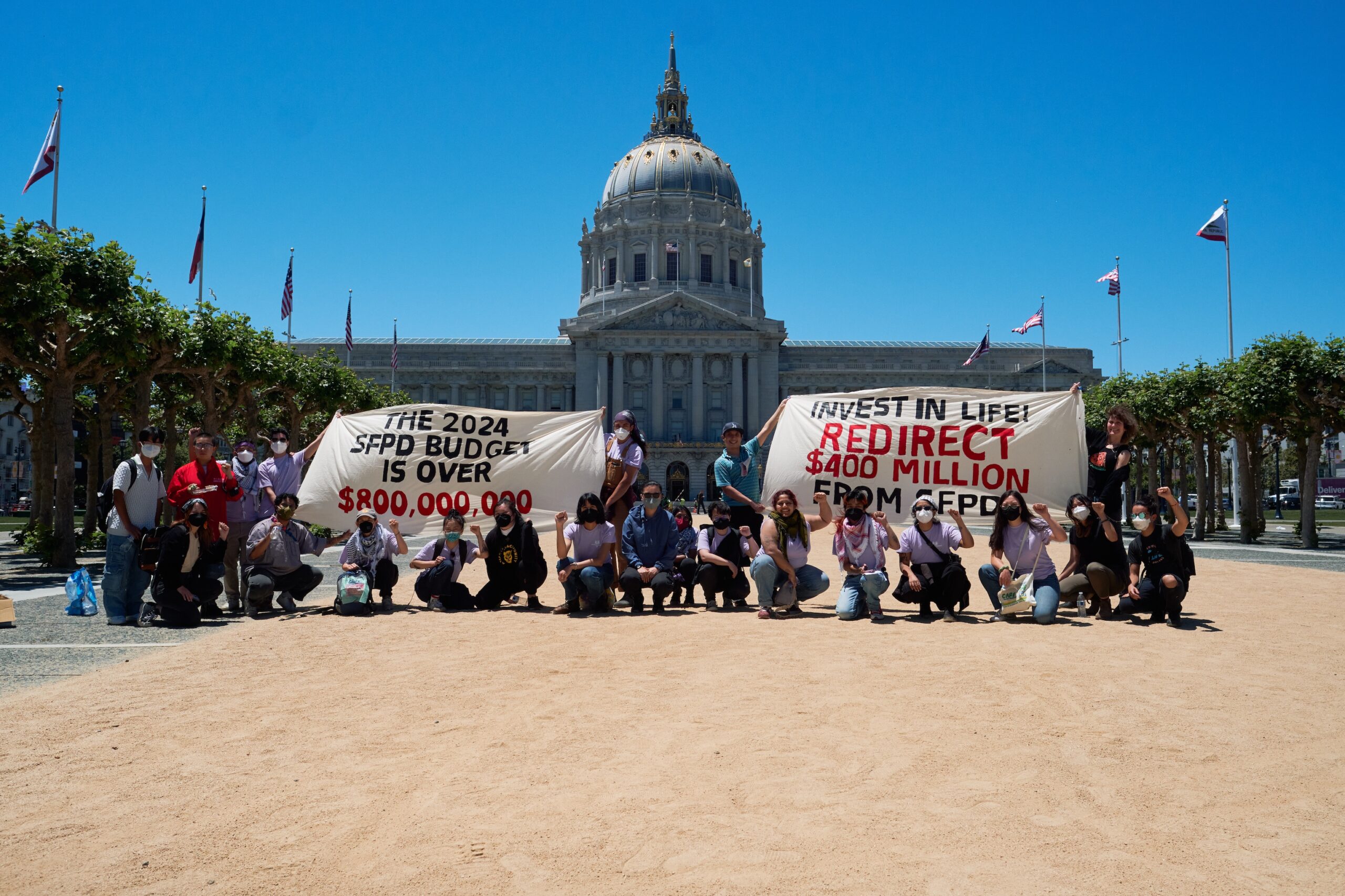 Image Description: A group of masked trans and queer Asian and Pacific Islander people hold a large purple banner that reads: Police Out of Pride. There is a black banner to the left that reads BLACK LIVES MATTER in multiple different languages.