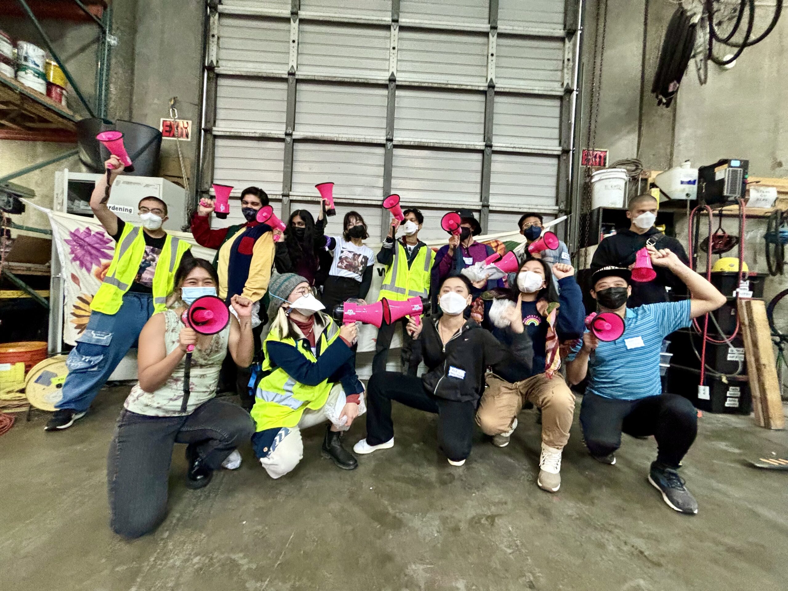 Thirteen queer and trans APIs pose in two rows front of a large warehouse door. Each person is masked, holding a hot pink megaphone and has a fist up.